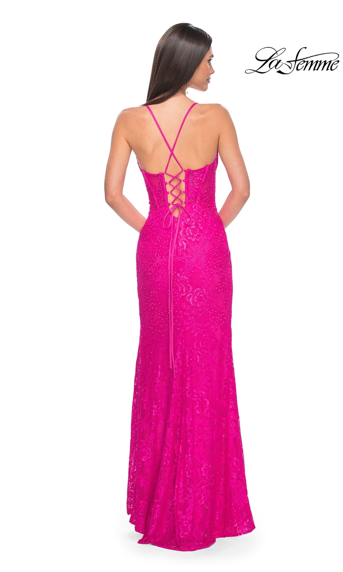 A woman stands facing away, wearing the La Femme Floral-Lace Long Corset Prom Dress 32248 in bright pink, featuring crisscrossed straps, a lace-up back, and a corset-style bodice.